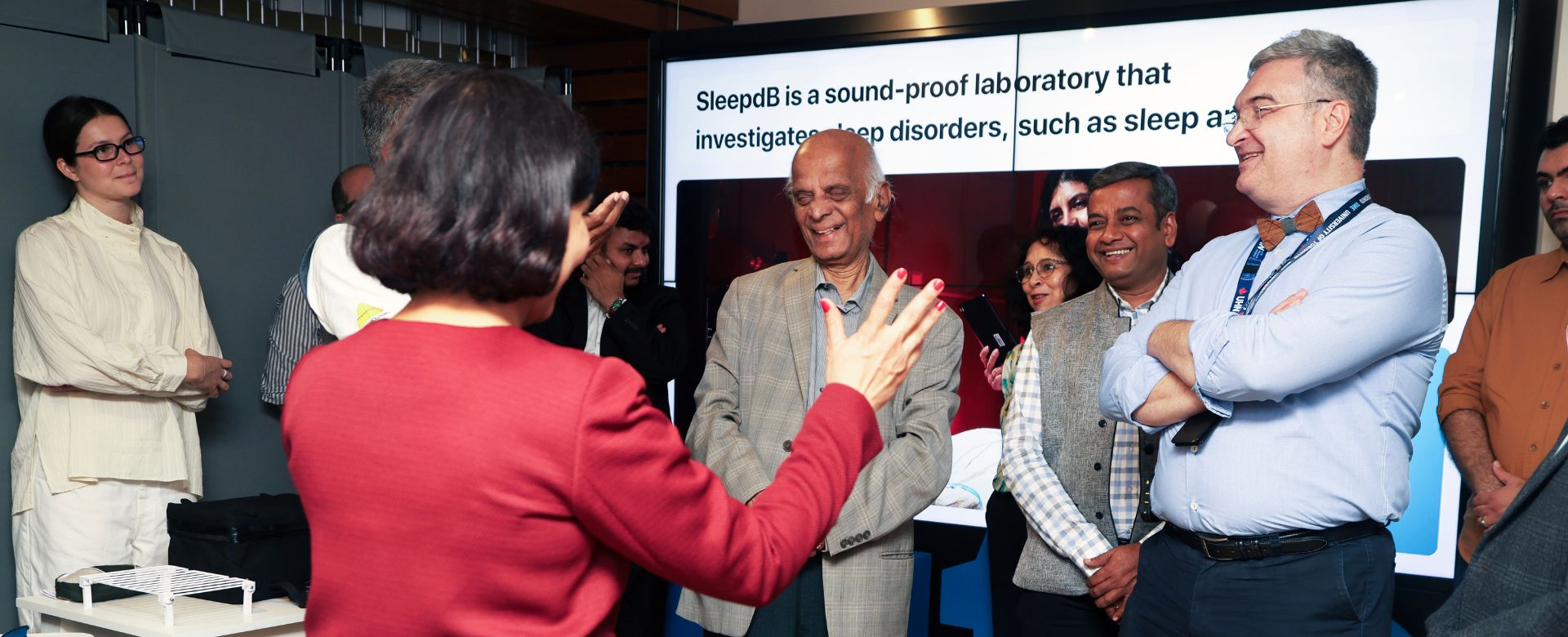 A group of people stand and listen attentively to a woman making a presentation 