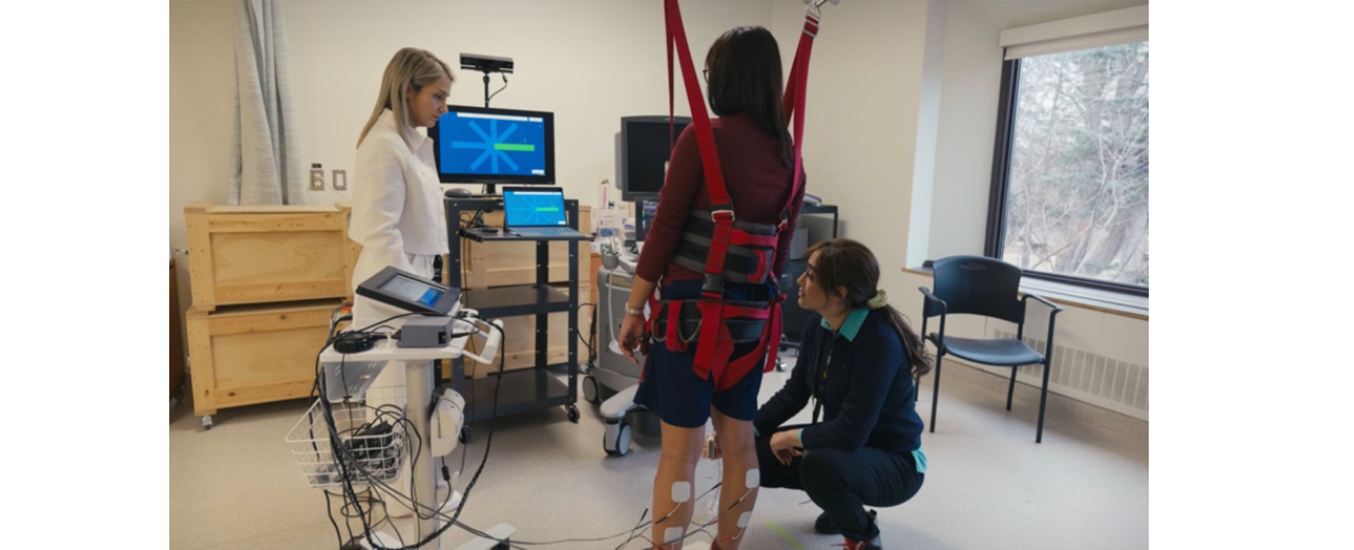 A woman standing and attached to a harness looks at a computer while two scientists watch her in a hospital room. 