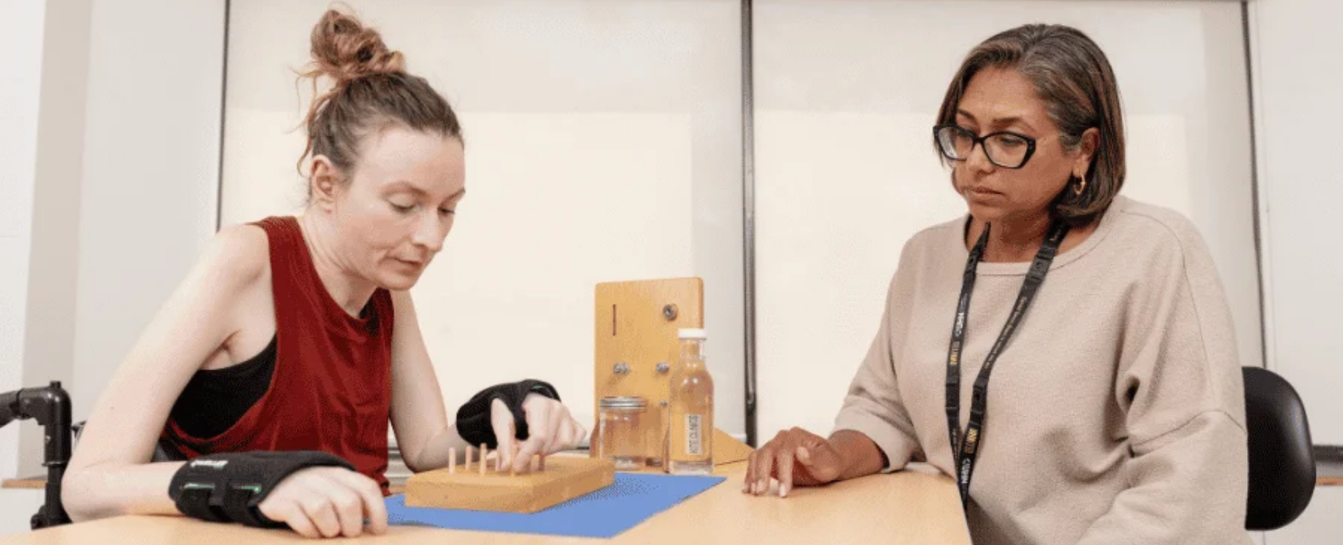 A woman in a wheelchair puts a peg into a board while a clinician looks on. 