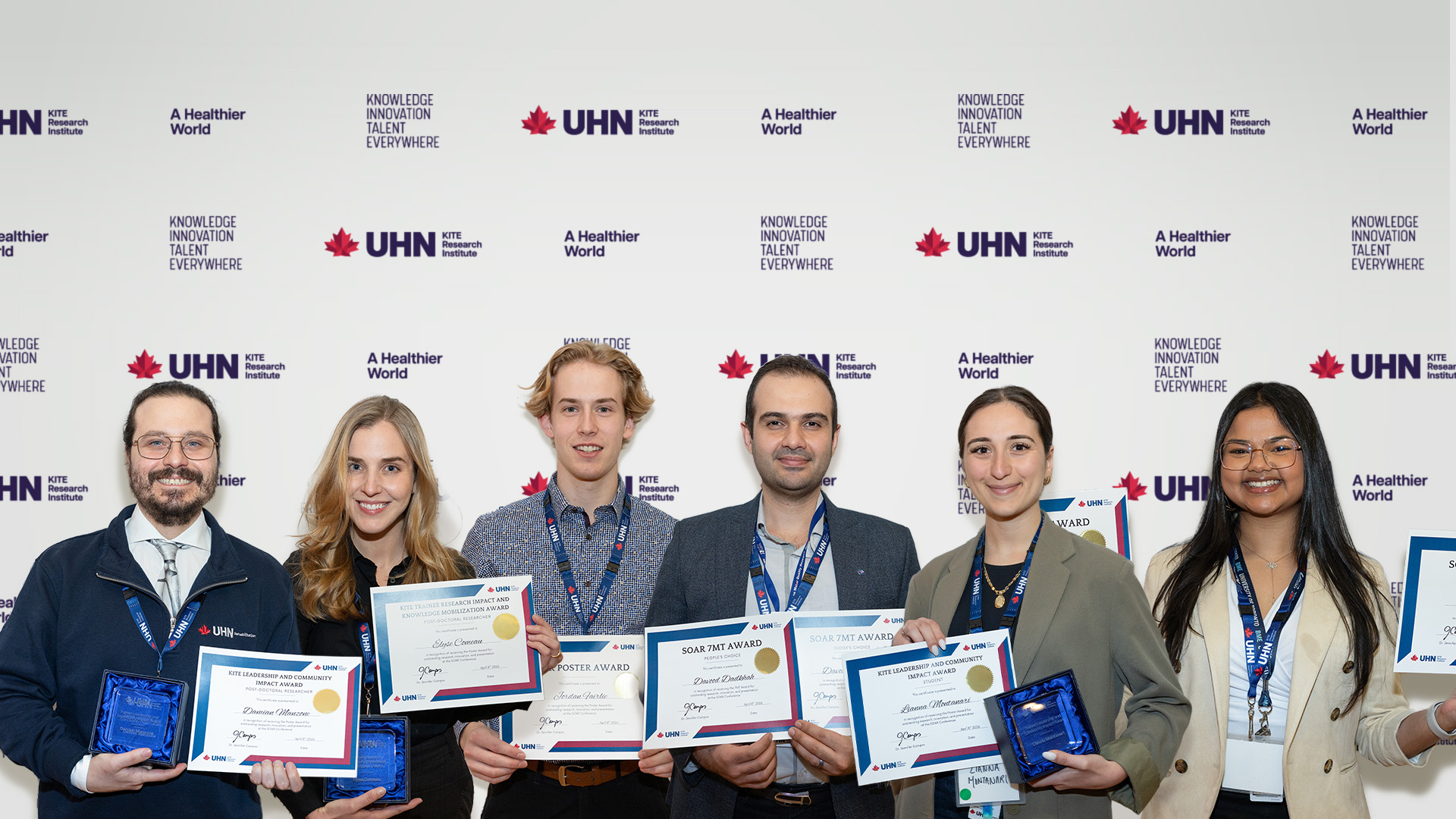 A photo with six trainee award winners holding paper awards standing in front of a backgrop that says UHN's KITE Research Institute. 