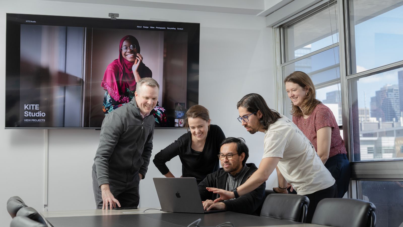 Five people gather around a computer screen in a bright boardroom.