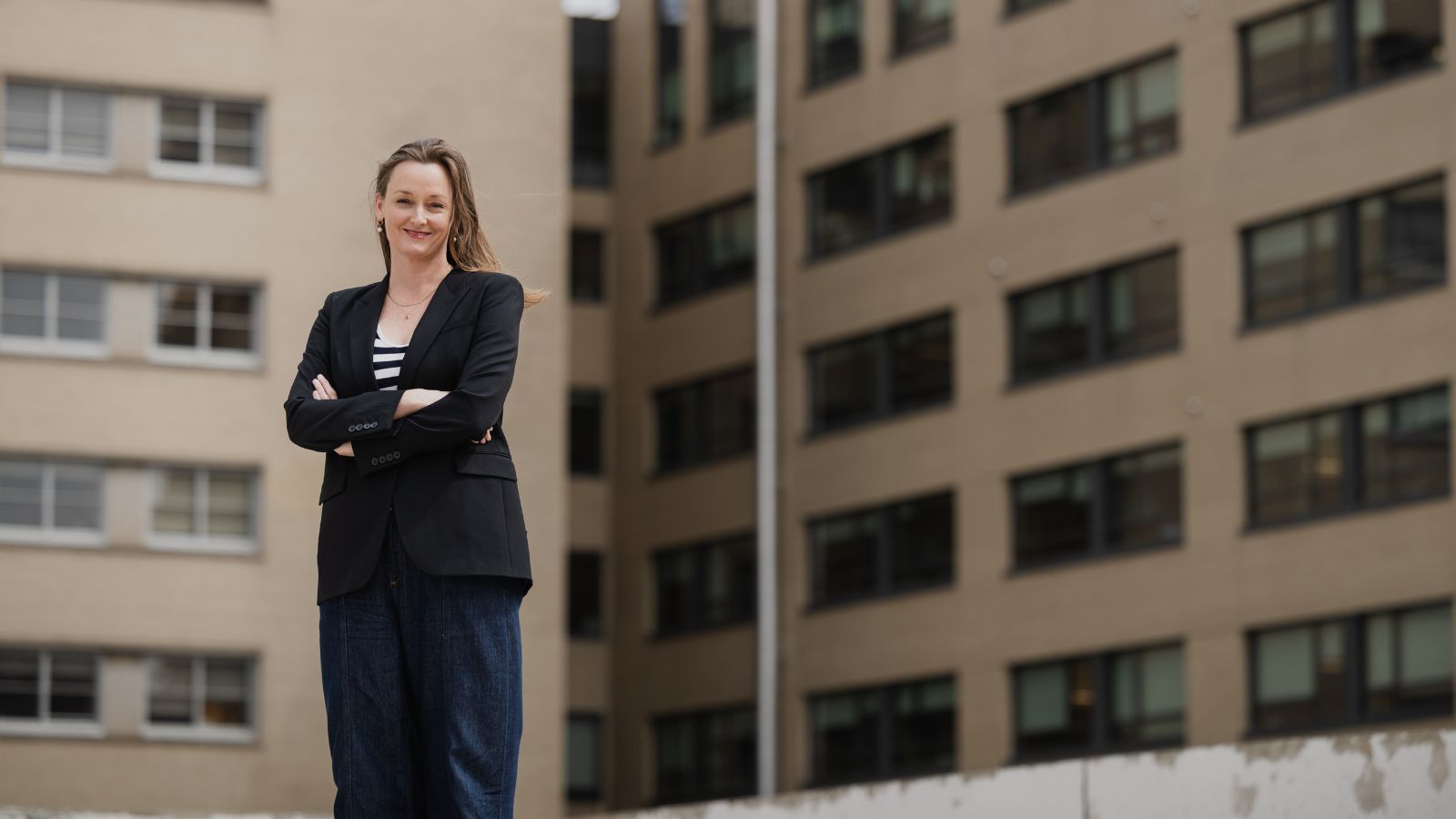 A woman with long hair in a blazer stands with her arms crossed in front of the side of a building with many windows behind her. 