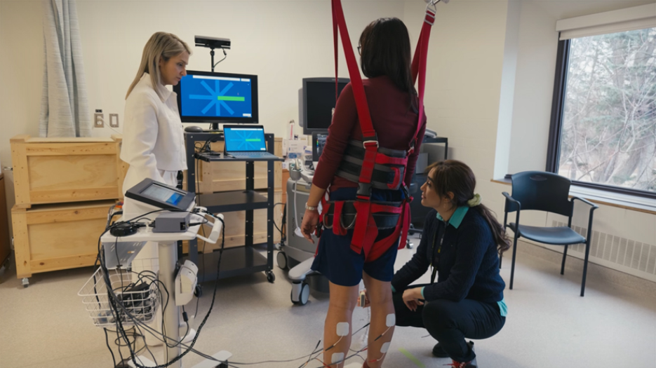 A woman stands in a harness in front of a computer with wires attached to her while two scientists stand next to her and look on. 